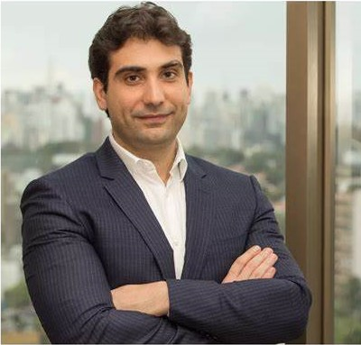Retrato de Gabriel Galípolo, de braços cruzados e paletó azul-marinho, com a paisagem urbana de São Paulo ao fundo.