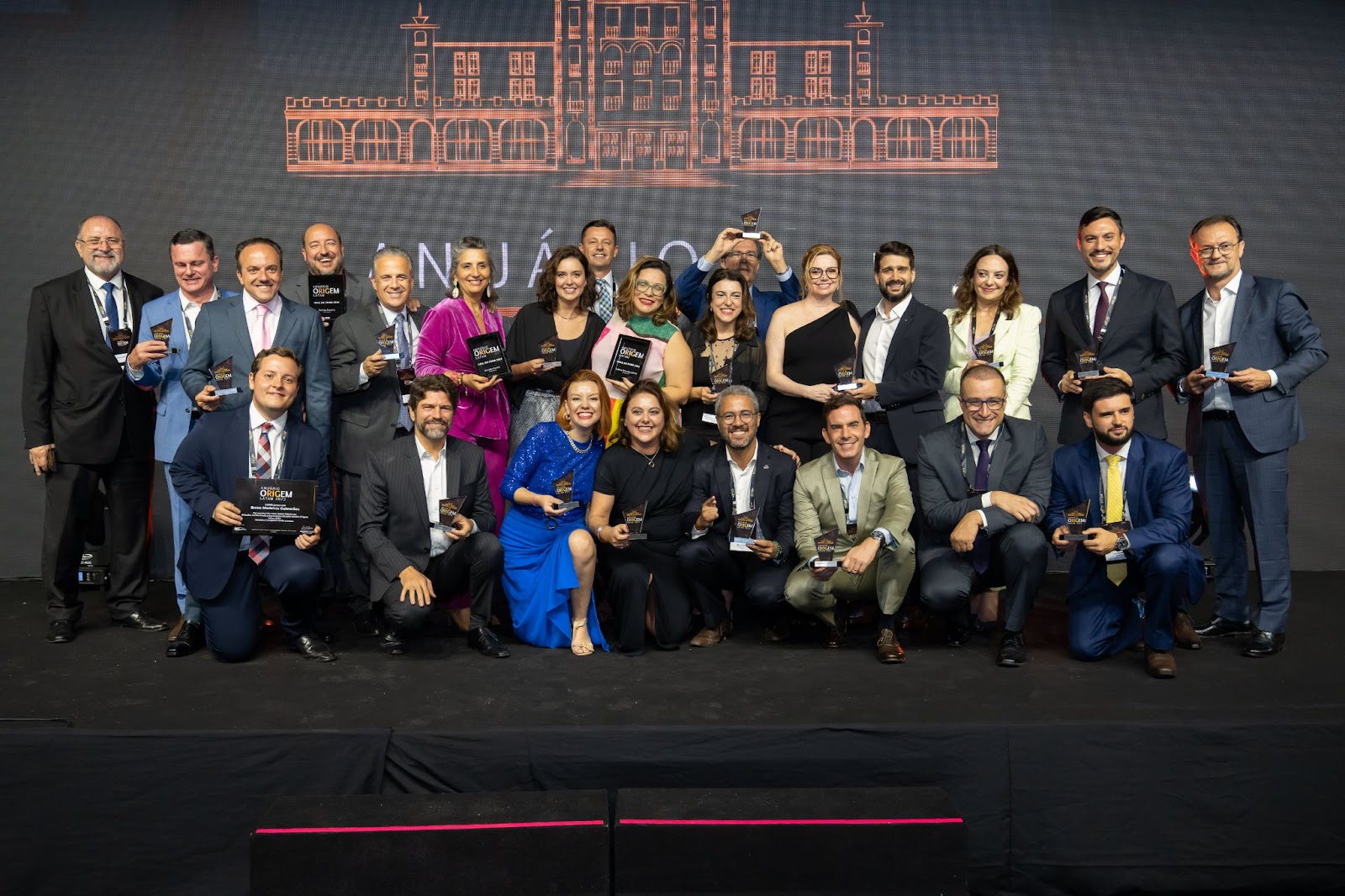 Foto de grupo dos premiados do Anuário ORIGEM 2023 no palco, segurando seus troféus.