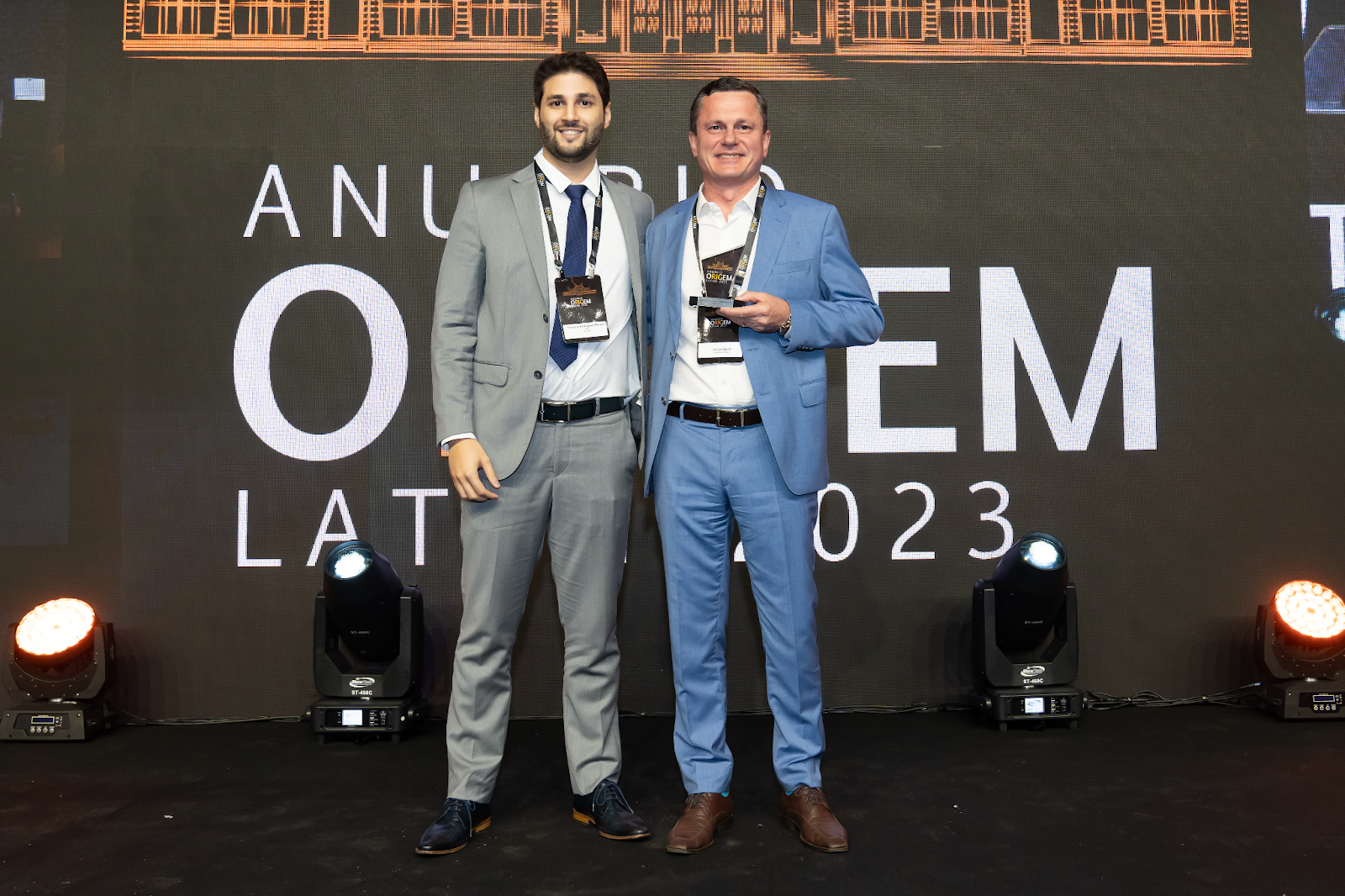Dois representantes da Nomos posando com o troféu no palco do Anuário ORIGEM LATAM 2023.